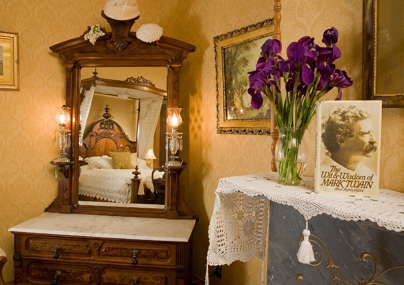 A vintage dresser with a mirror reflecting a four-poster bed, a vase of purple flowers, and framed a