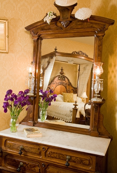 A vintage dresser with a mirror reflecting a four-poster bed, a vase of purple flowers, and framed a