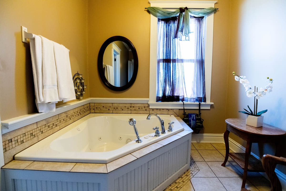This elegant bathroom features a corner jetted tub, a round mirror, and a window with sheer dark blu