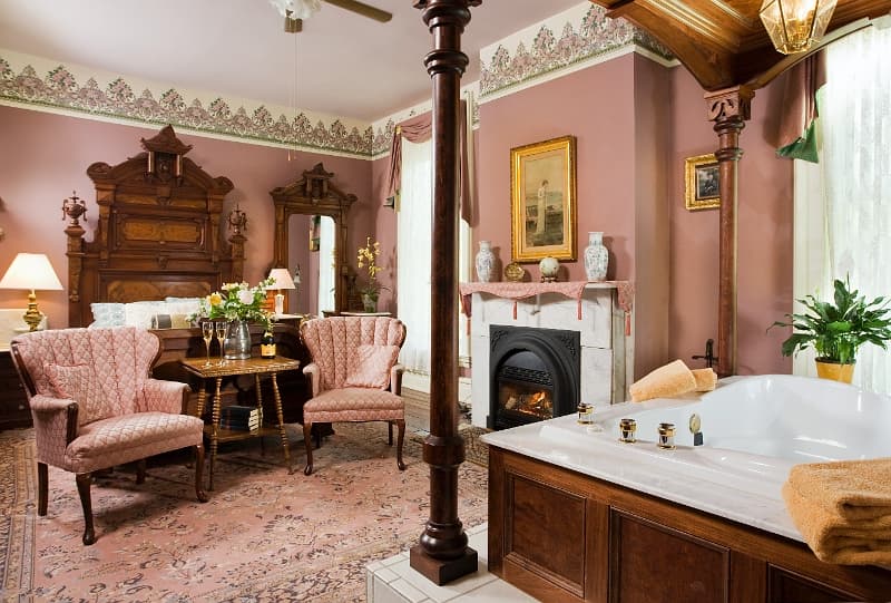 A spacious bedroom with a four-poster bed, a cozy fireplace, plush armchairs, and a luxurious jacuzz