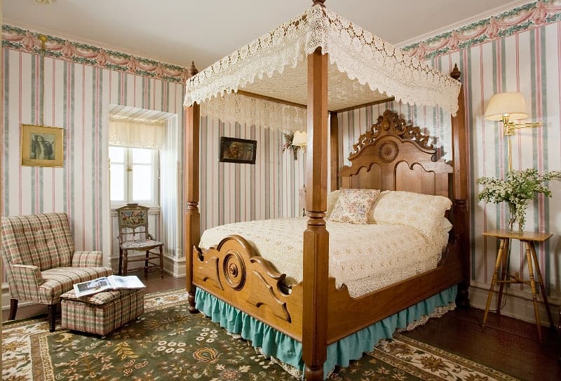 A bedroom with a large four-poster bed, a sitting area with an armchair and ottoman, and striped wal