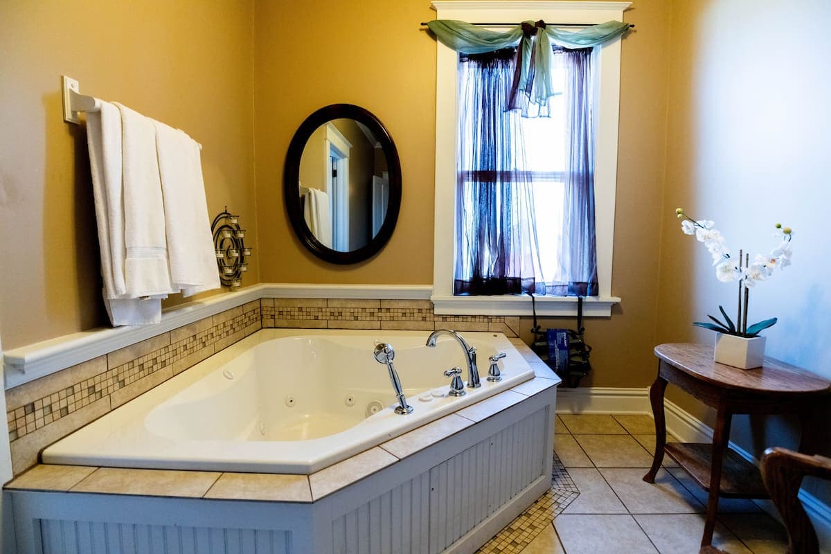 This elegant bathroom features a corner jetted tub, a round mirror, and a window with sheer dark blu