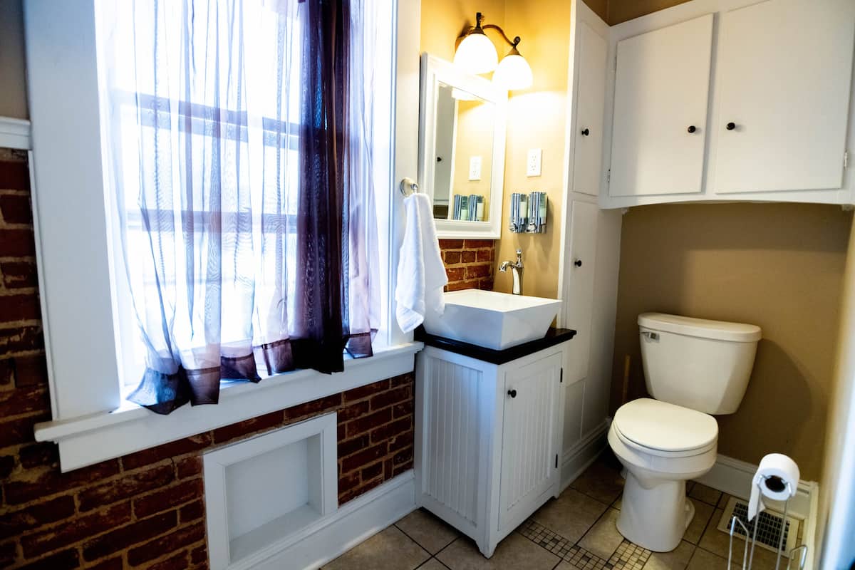 This cozy bathroom features exposed brick, a white vessel sink on a vanity, a toilet, built-in stora
