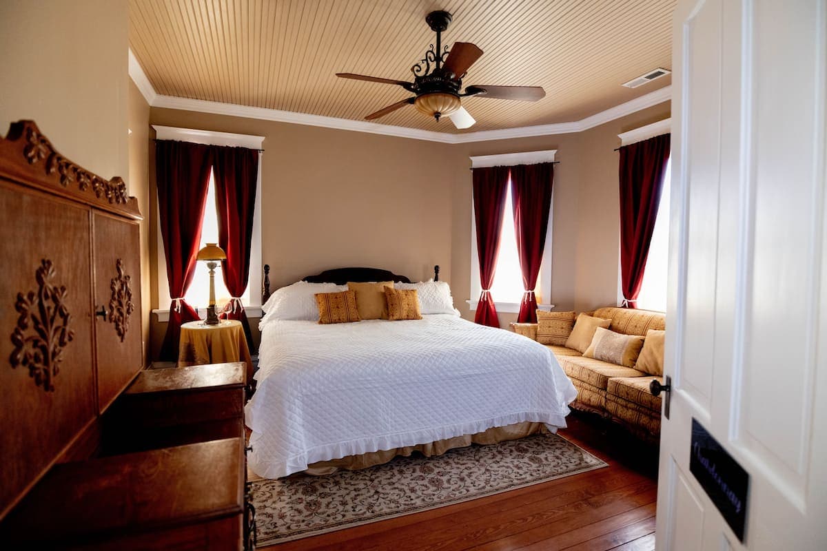 A classic bedroom featuring a large bed with white linens, a dark wood headboard, red curtains, a c