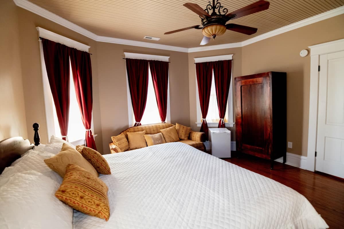 A spacious bedroom with a large bed, yellow accent pillows, a dark wood armoire, a sofa, a ceiling f