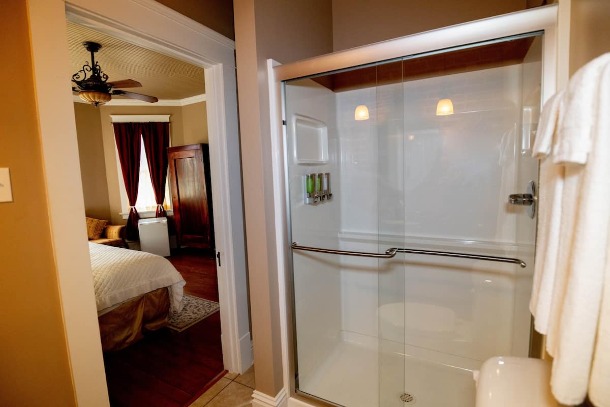 A bright bathroom with a glass-enclosed shower, a toilet, and white towels, with a view into an adj