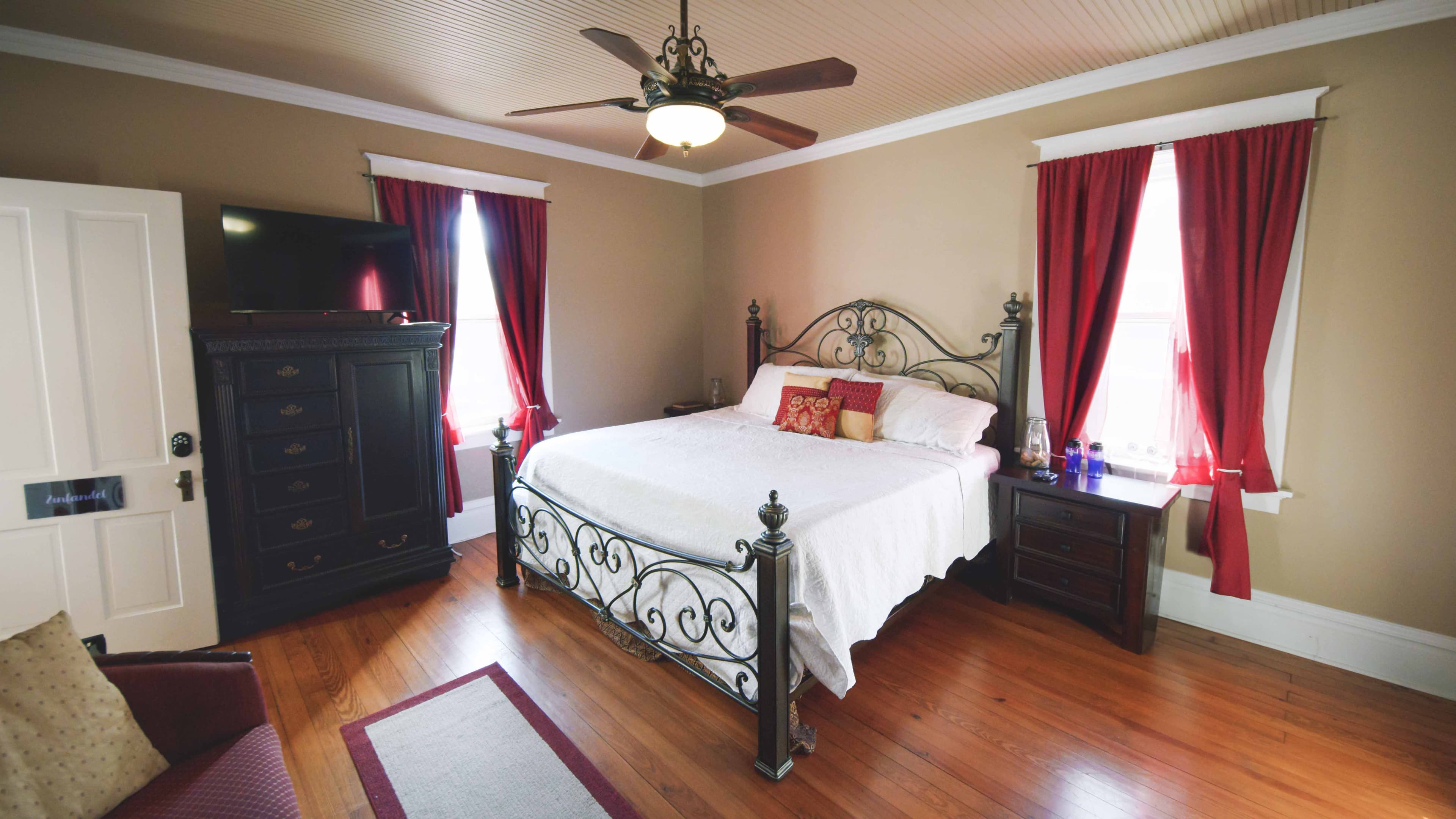 This cozy bedroom features a metal-frame bed with white bedding, dark wood furniture, rich red curt