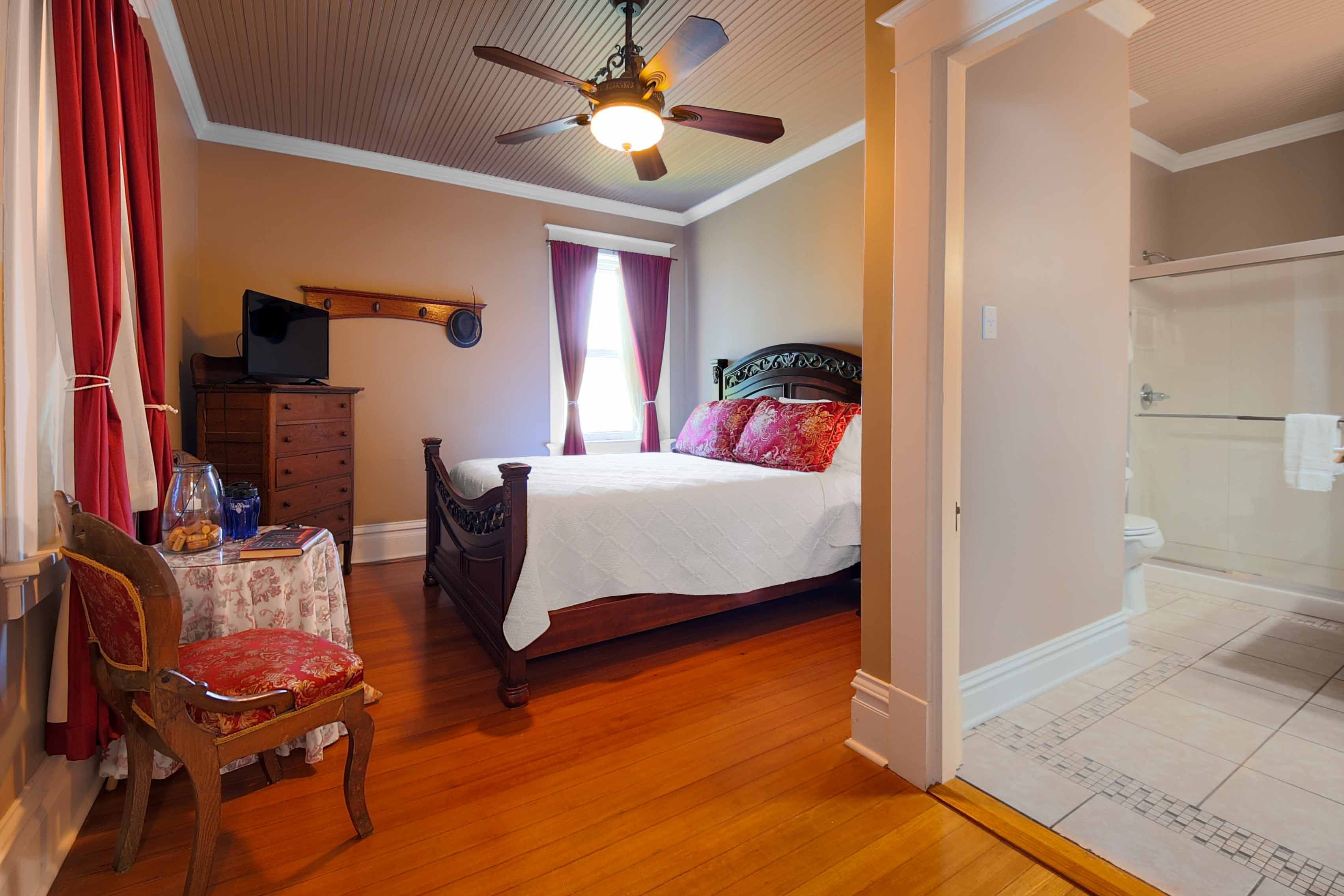 A charming bedroom featuring a bed with colorful pillows, hardwood floors, a ceiling fan, red drape