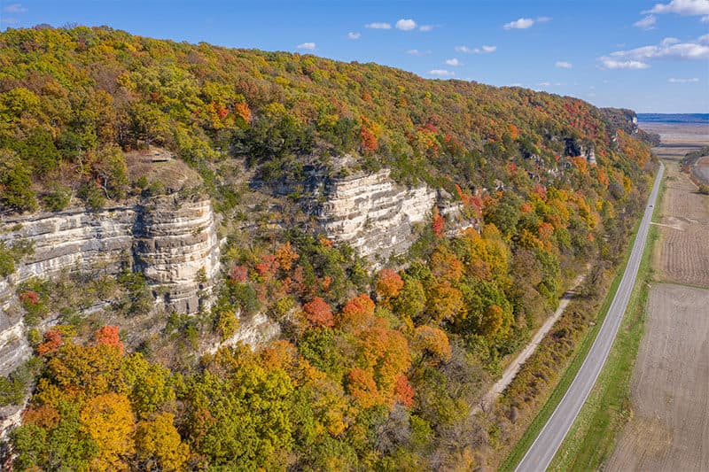 Aerial view of a colorful autumn forest along a rocky cliff, with a road running beside it.