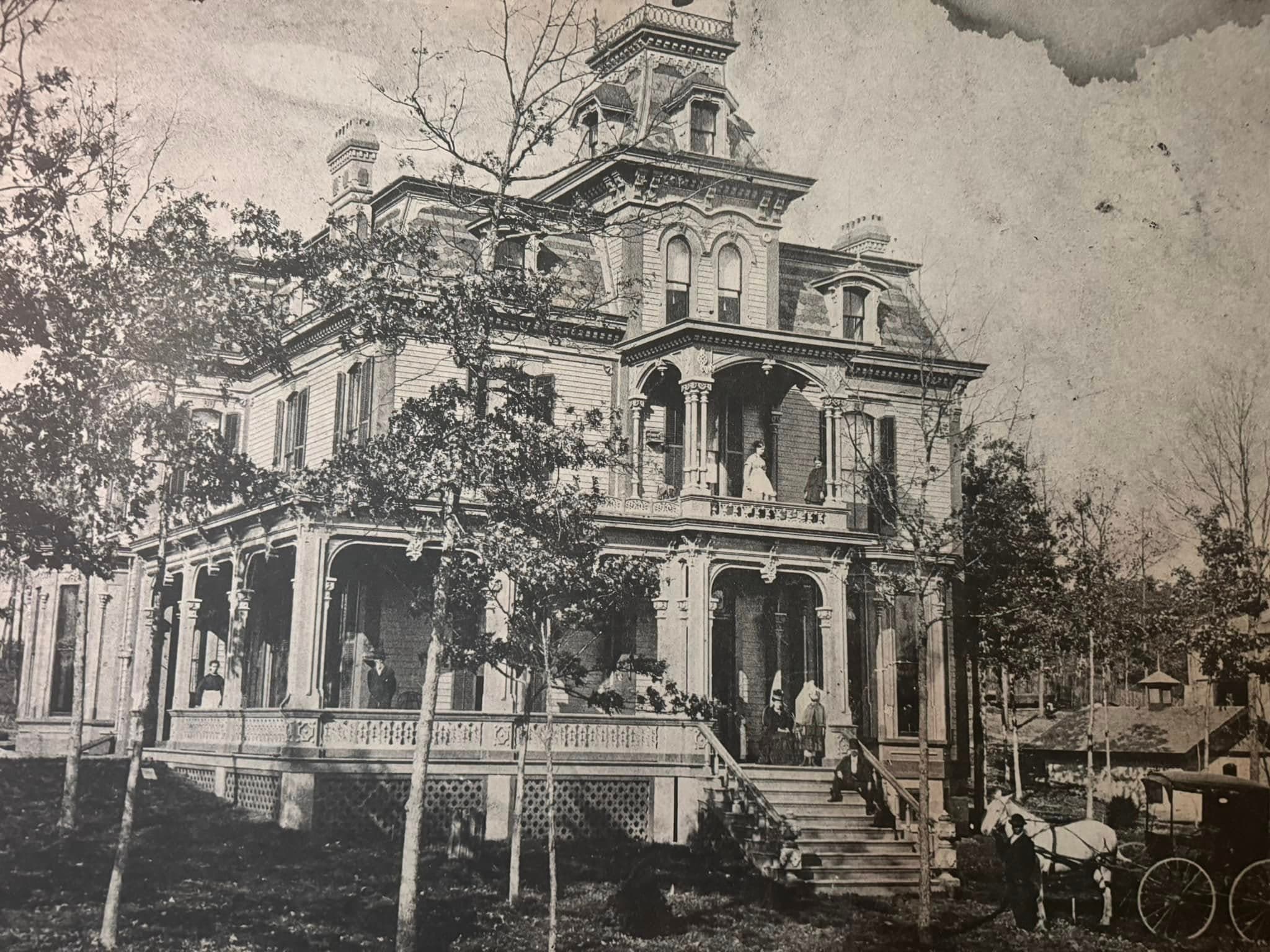 A historic Victorian house surrounded by trees, with people and a horse-drawn carriage in view.