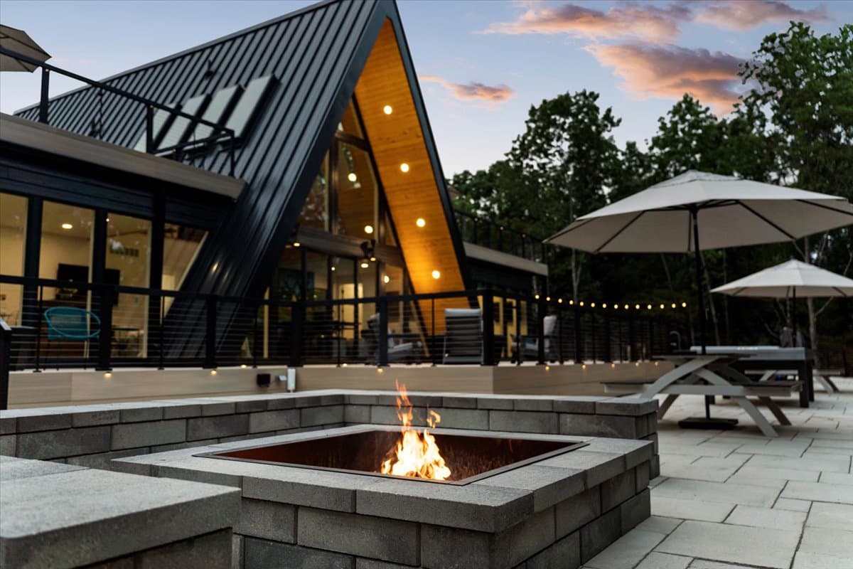 A modern cabin with a triangular roof overlooks a stone fire pit and patio area under a twilight sky.