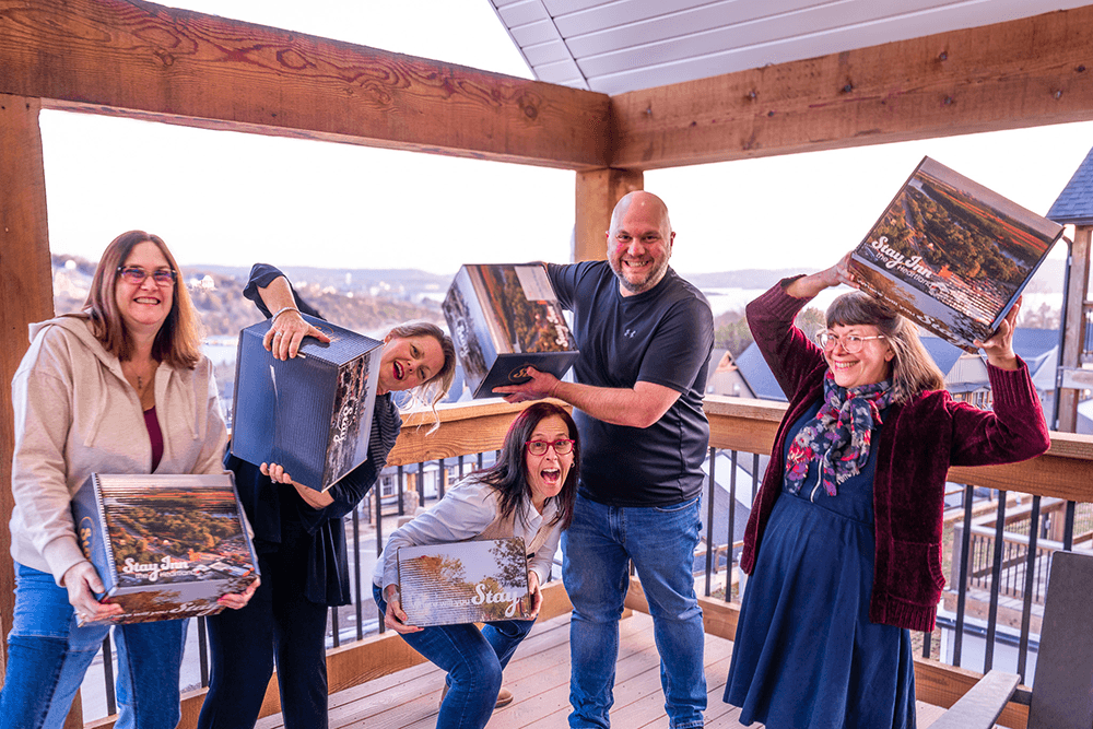 Five people are joyfully posing with large boxes on a wooden balcony overlooking a scenic view.