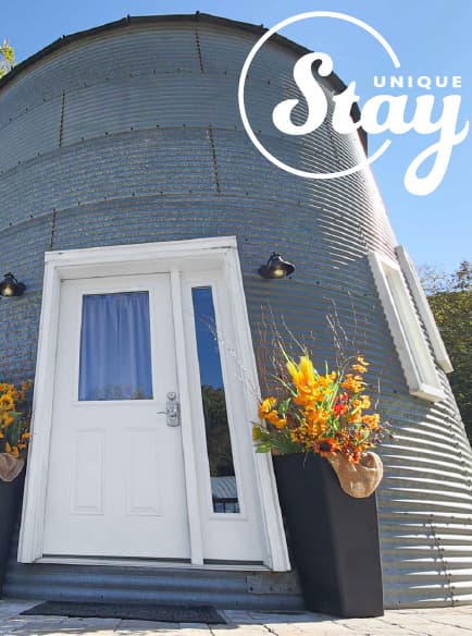 A unique, cylindrical metal building features a white door and vibrant flower arrangements at the entrance.