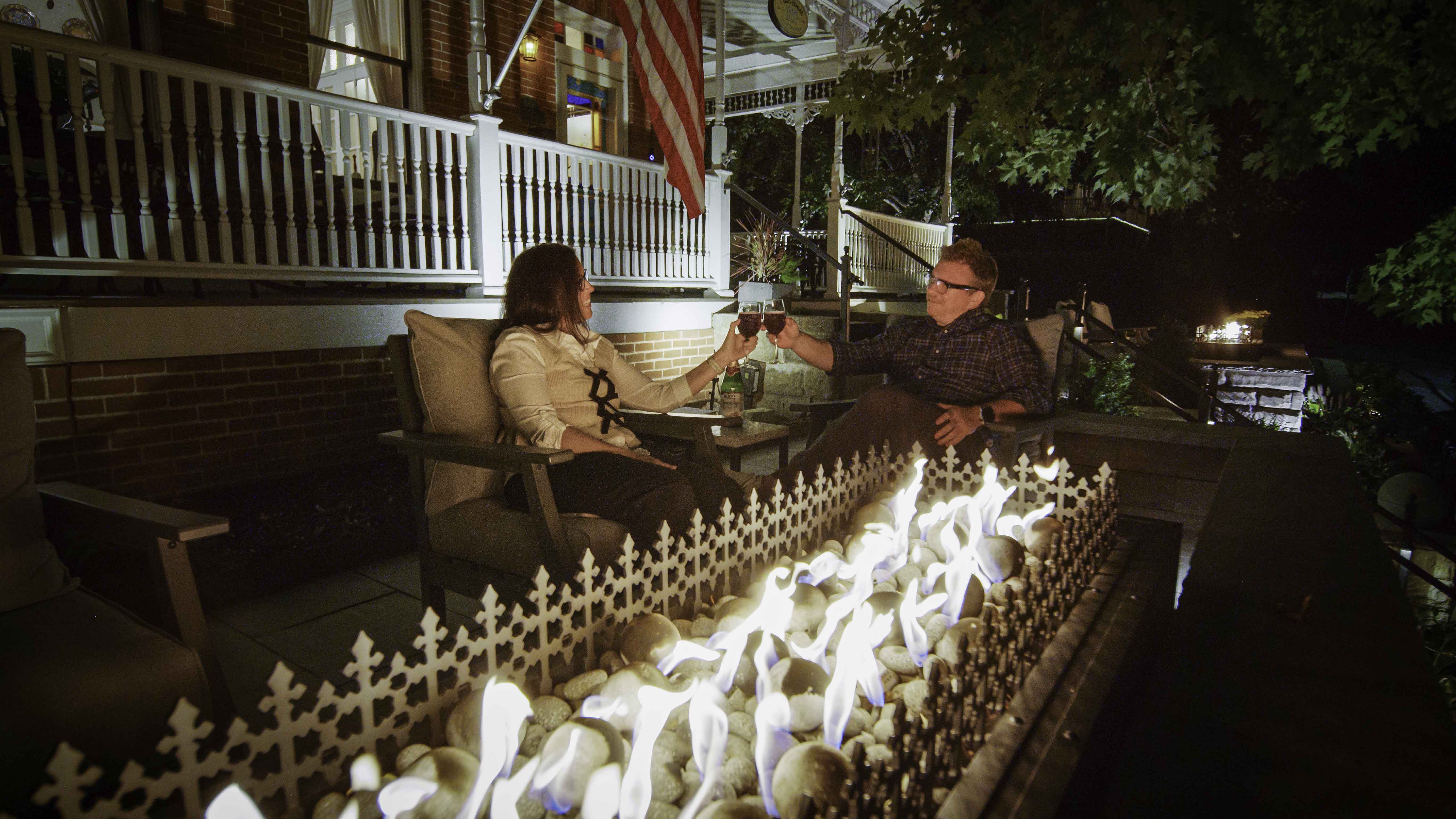 Two people toast drinks by a glowing fire feature on a cozy porch at night.