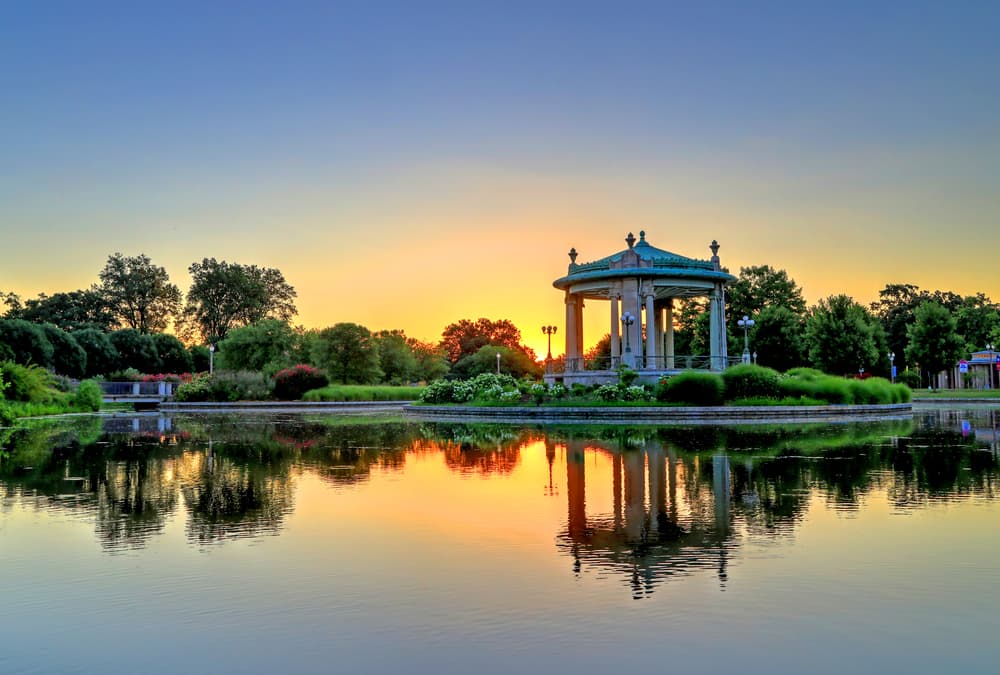 A gazebo reflects in a serene pond at sunset, surrounded by lush greenery.