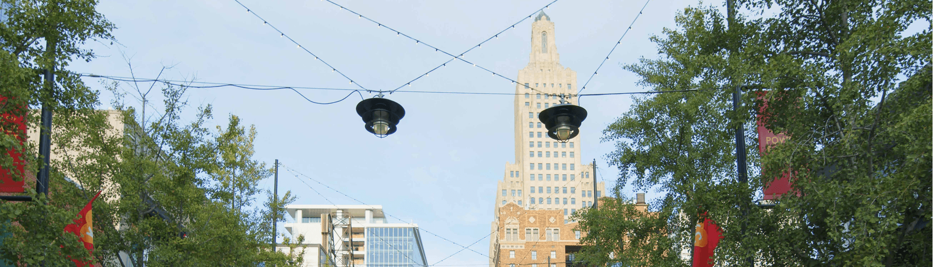 A city street lined with trees, decorative lights, and a prominent skyscraper in the background.