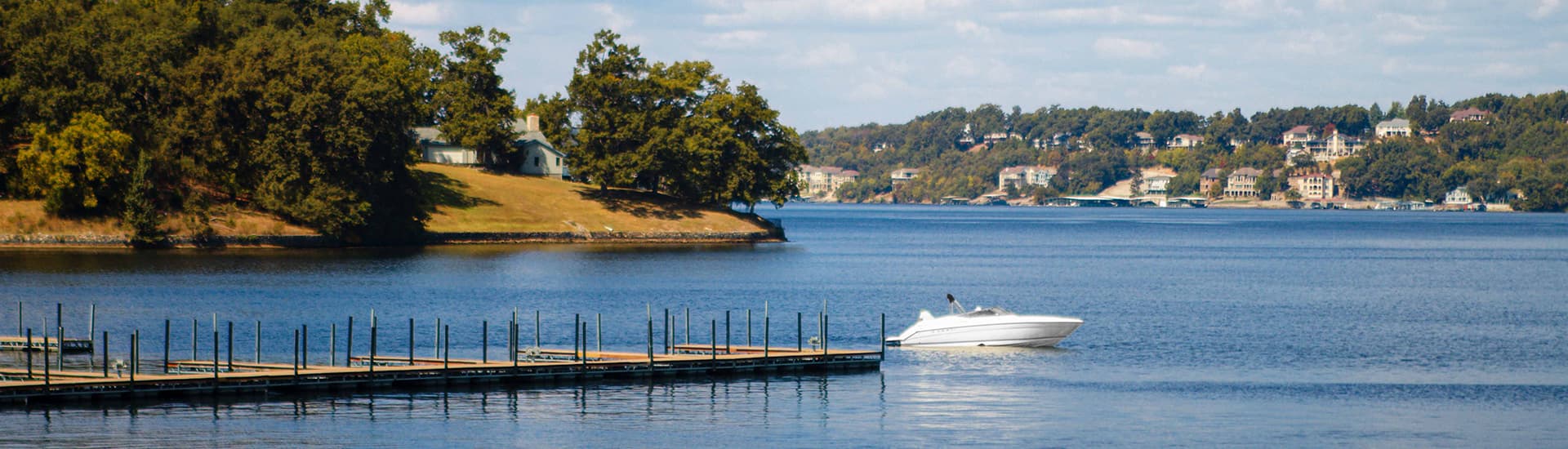 A boat glides across a calm lake near a wooded shoreline and distant homes.