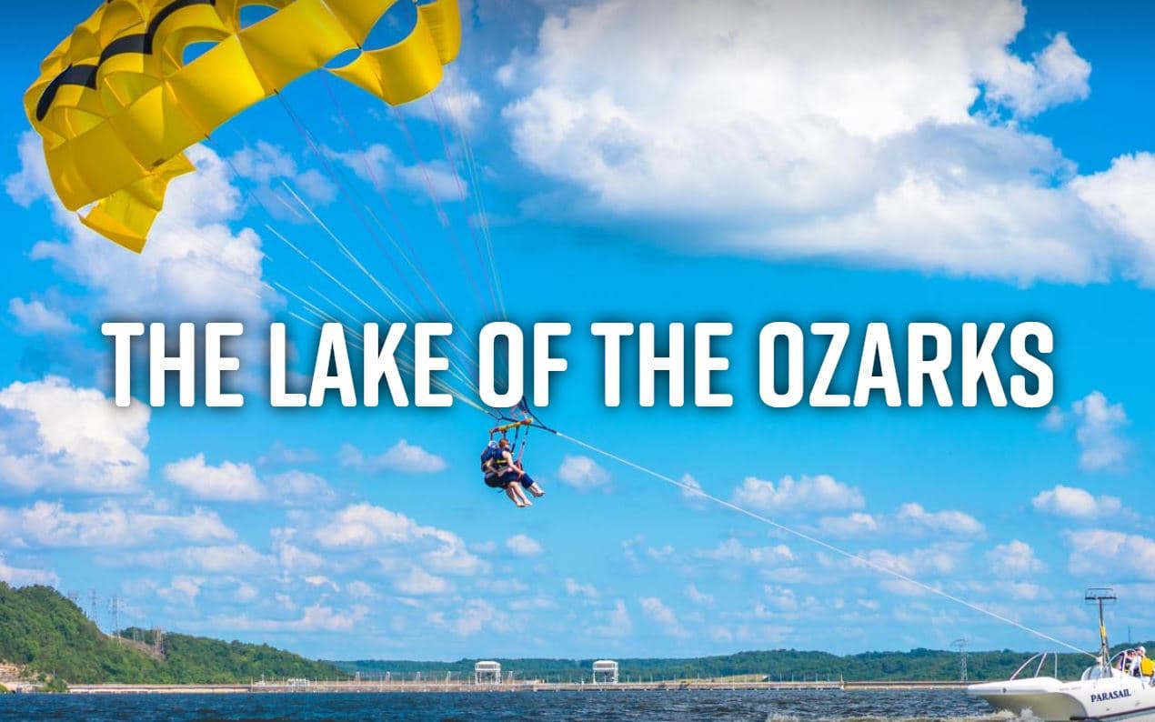 A person paraglides over the Lake of the Ozarks under a bright blue sky.