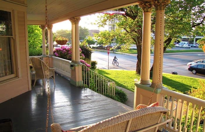A sunny porch view with swinging chairs, flowers, and a cyclist passing by.