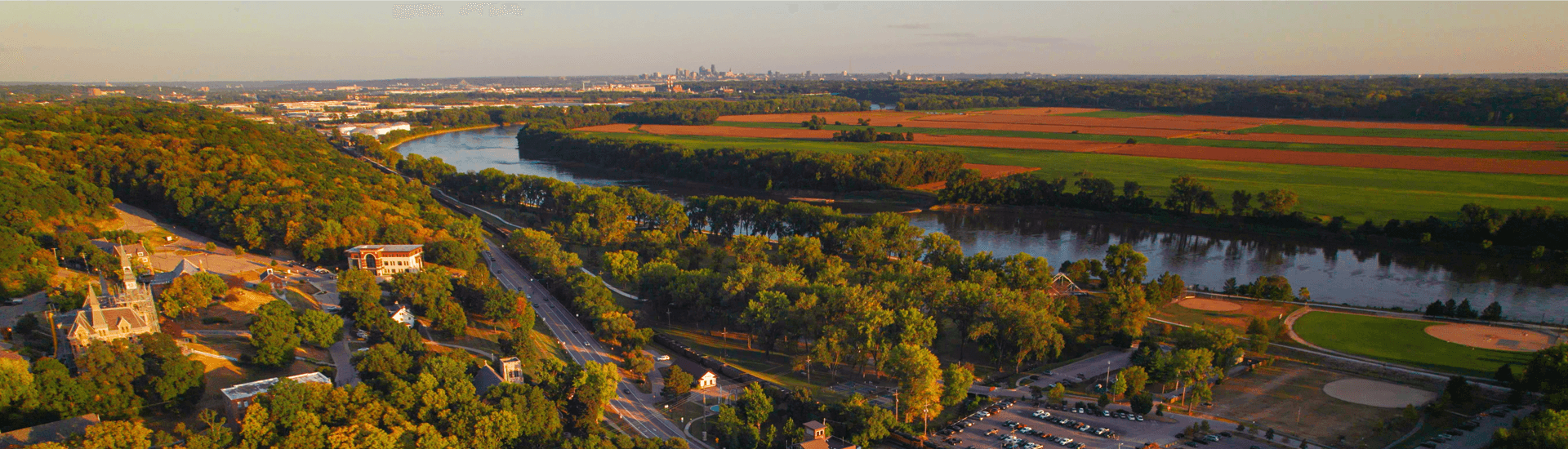 A scenic aerial view of a river winding through green forests and fields, with a distant city skyline.