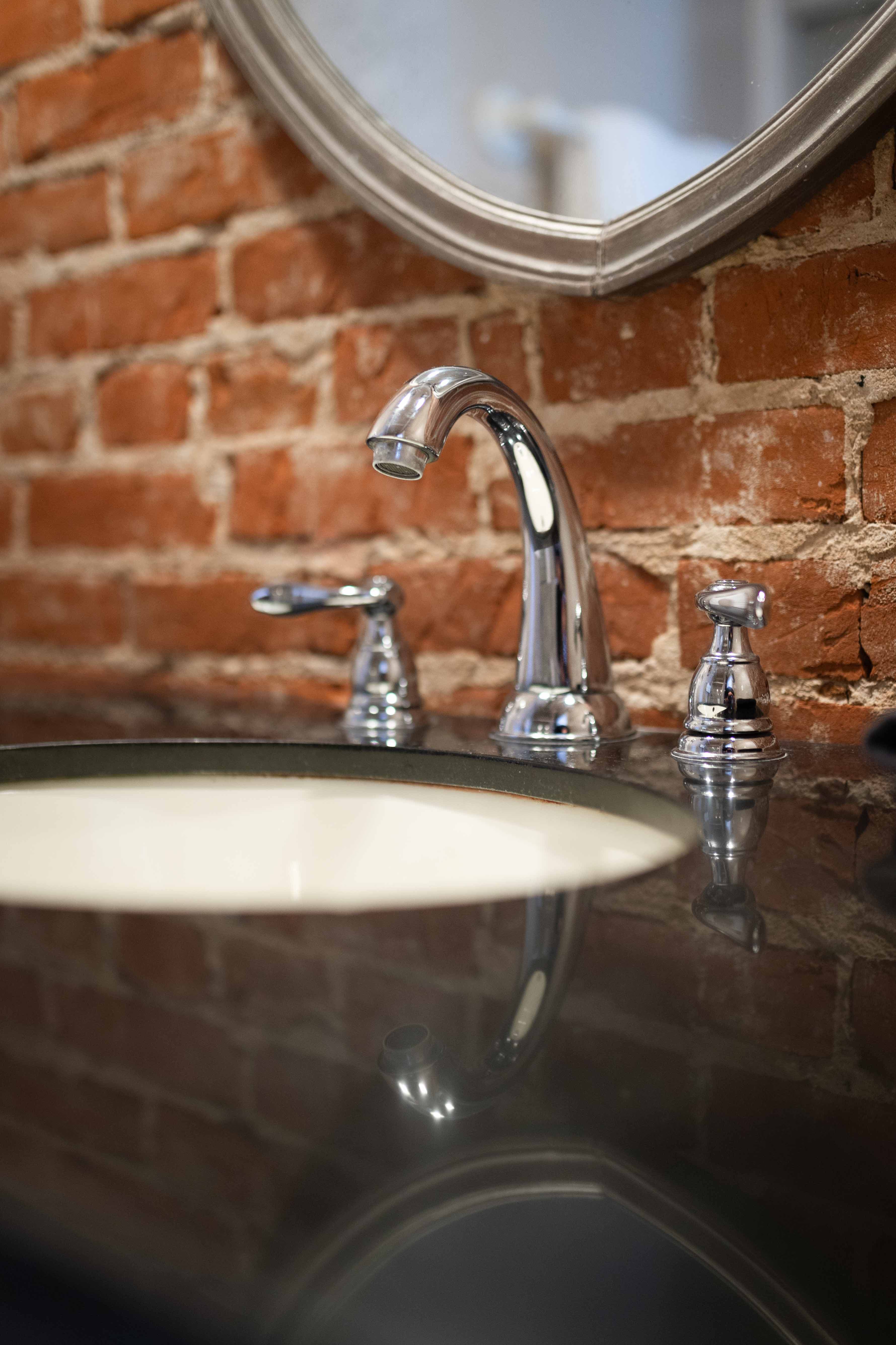 A close-up of a chrome faucet and sink reflecting against a rustic brick wall.