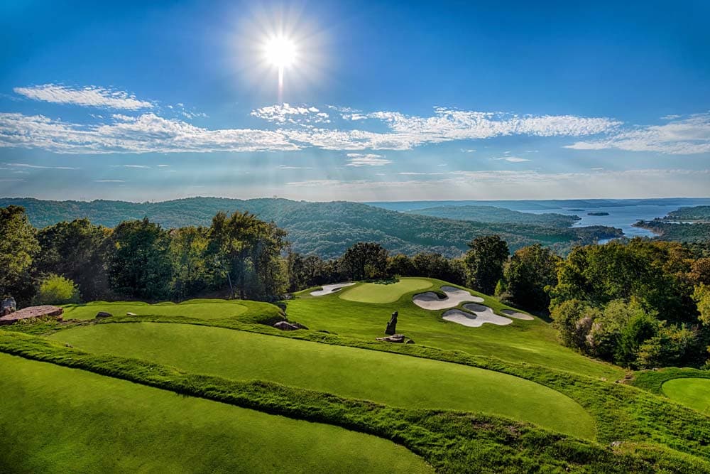 A scenic view of a golf course with rolling hills and a lake under a bright sun.