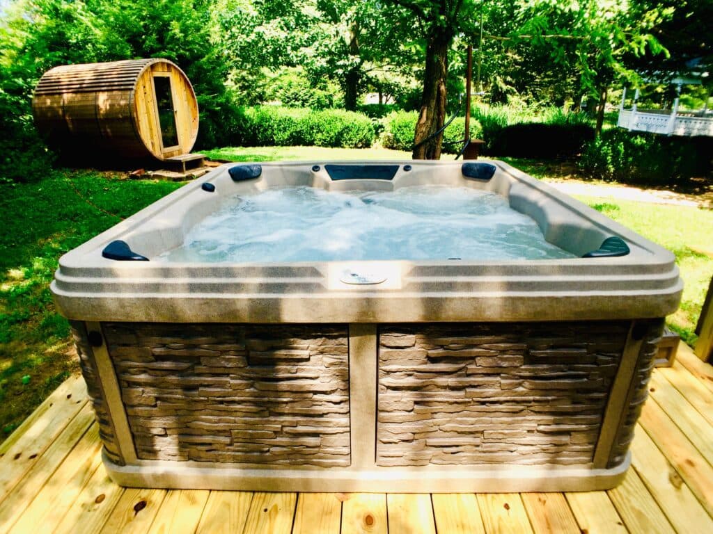 A hot tub on a wooden deck surrounded by greenery and a sauna in the background.