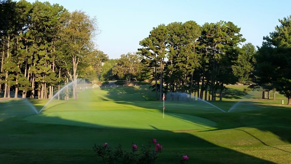 A lush green golf course with sprinklers watering the grass under a clear blue sky.