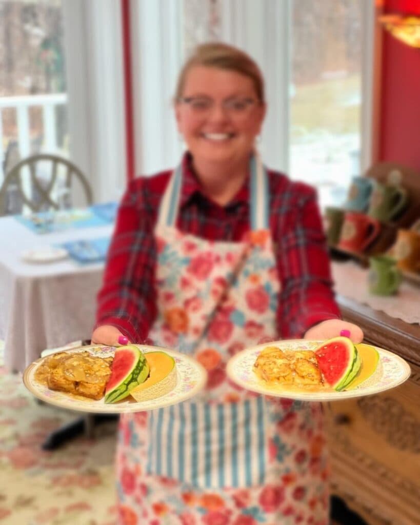 A smiling woman in a floral apron holds two plates of food with fruit garnishes.