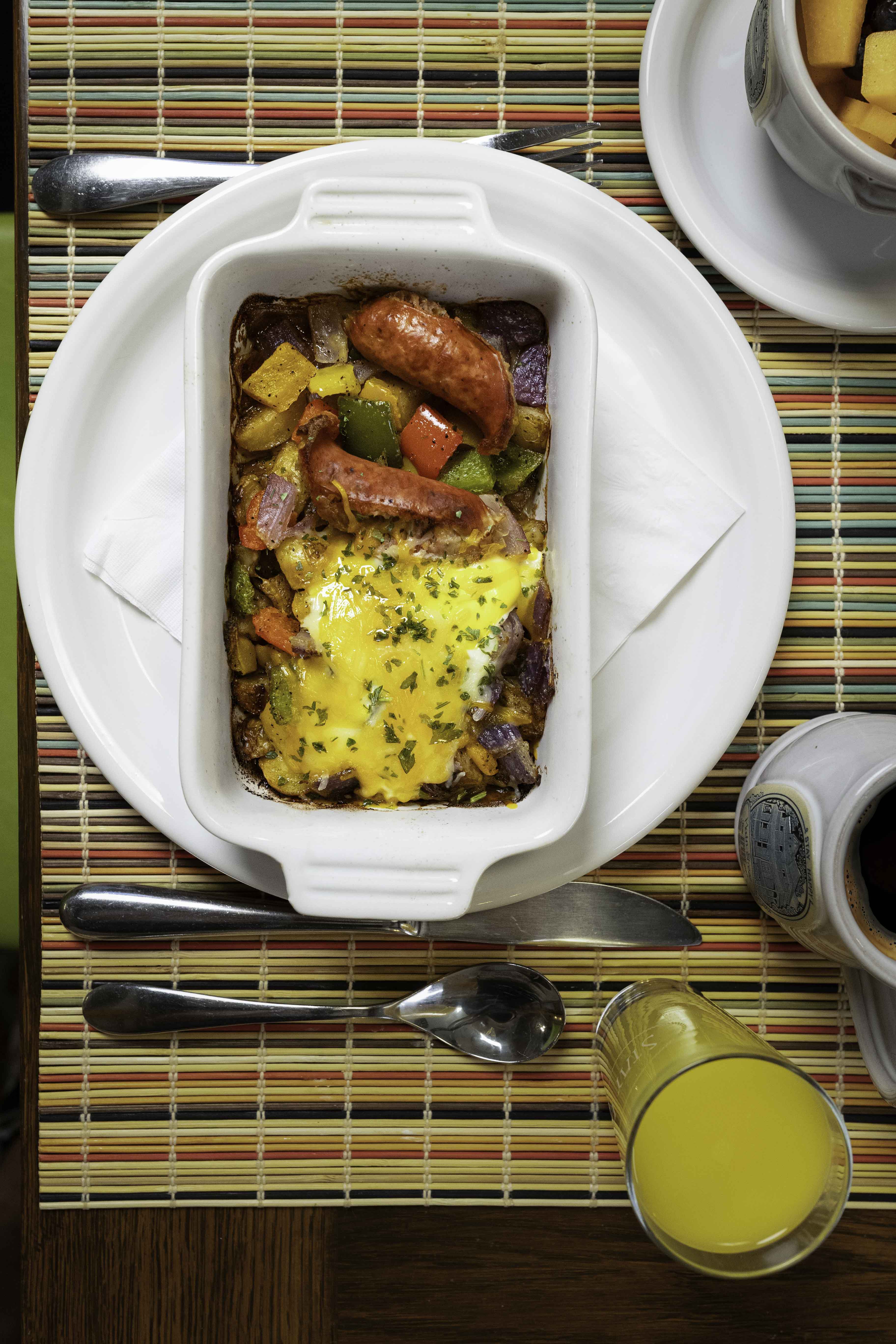 A baked dish with sausage, peppers, and melted cheese served on a colorful placemat, accompanied by a glass of orange juice and a cup of coffee.