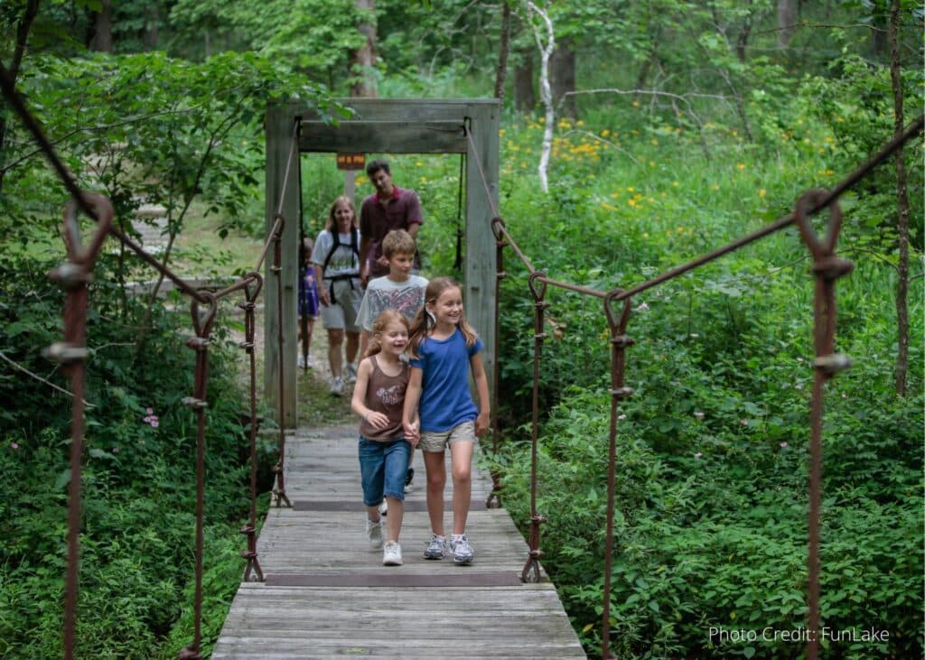 A group of children walks across a wooden bridge in a lush green forest.
