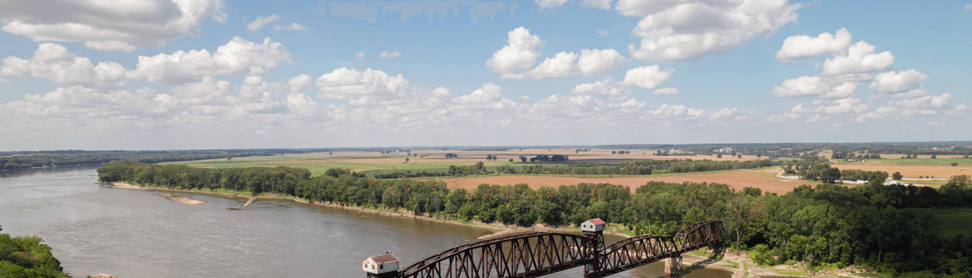 A panoramic view of a river winding through lush greenery and expansive farmland under a blue sky filled with clouds.