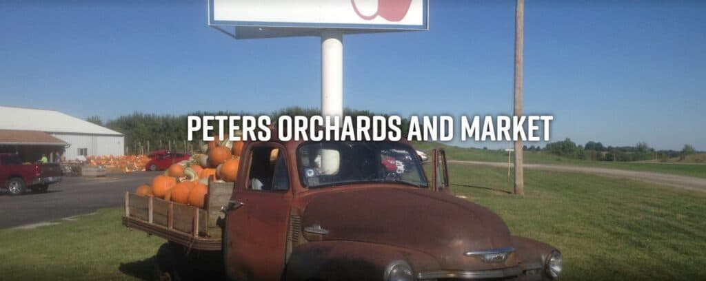 An old truck filled with pumpkins parked in front of a market sign that reads "Peters Orchards and Market."