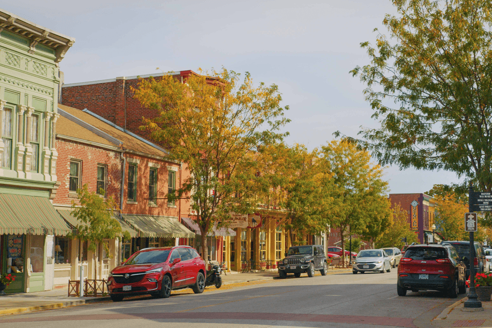 A charming main street lined with colorful autumn trees and historic buildings, featuring parked cars and small shops.