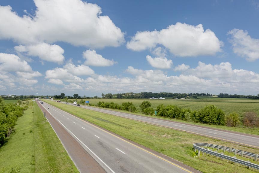 A wide view of a highway flanked by green fields and a blue sky with fluffy clouds. A wide view of a highway flanked by green fields and a blue sky with fluffy clouds.