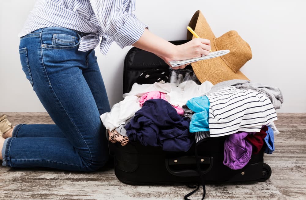 A person kneels beside a suitcase overflowing with clothes while taking notes. A person kneels beside a suitcase overflowing with clothes while taking notes.