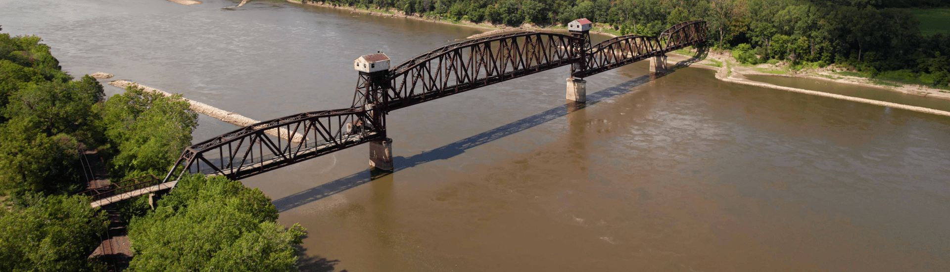 Aerial view of a rust-colored railway bridge over a river surrounded by green trees.