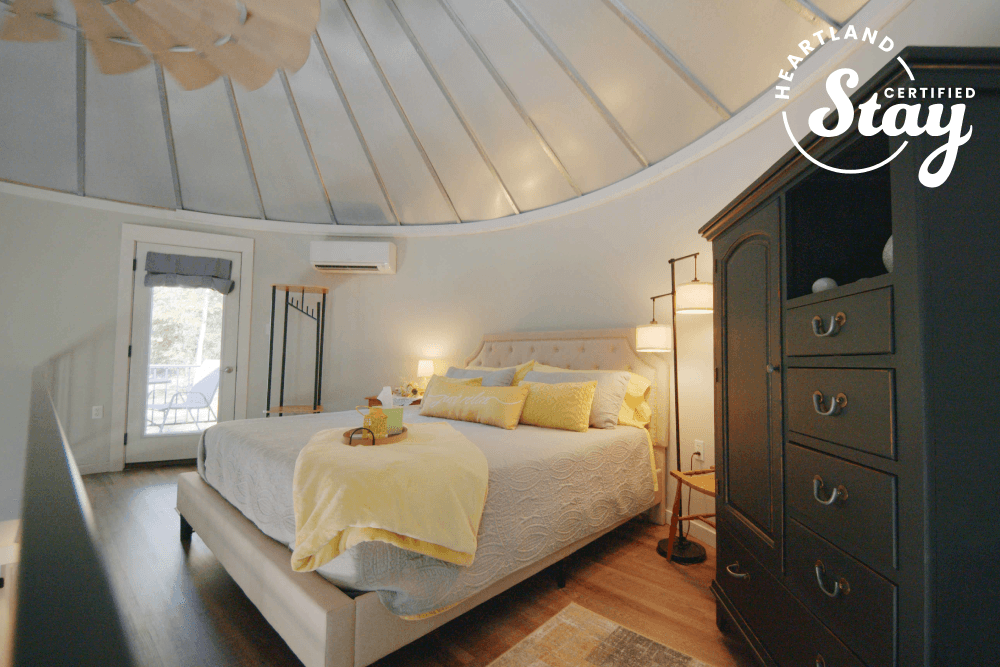 A cozy bedroom with a circular ceiling, featuring a large bed dressed in yellow accents, a dark wooden dresser, and natural light from a nearby door.