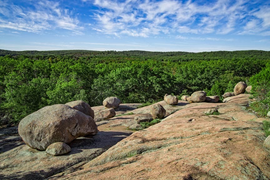 A rocky landscape with large boulders surrounded by lush green trees under a blue sky.