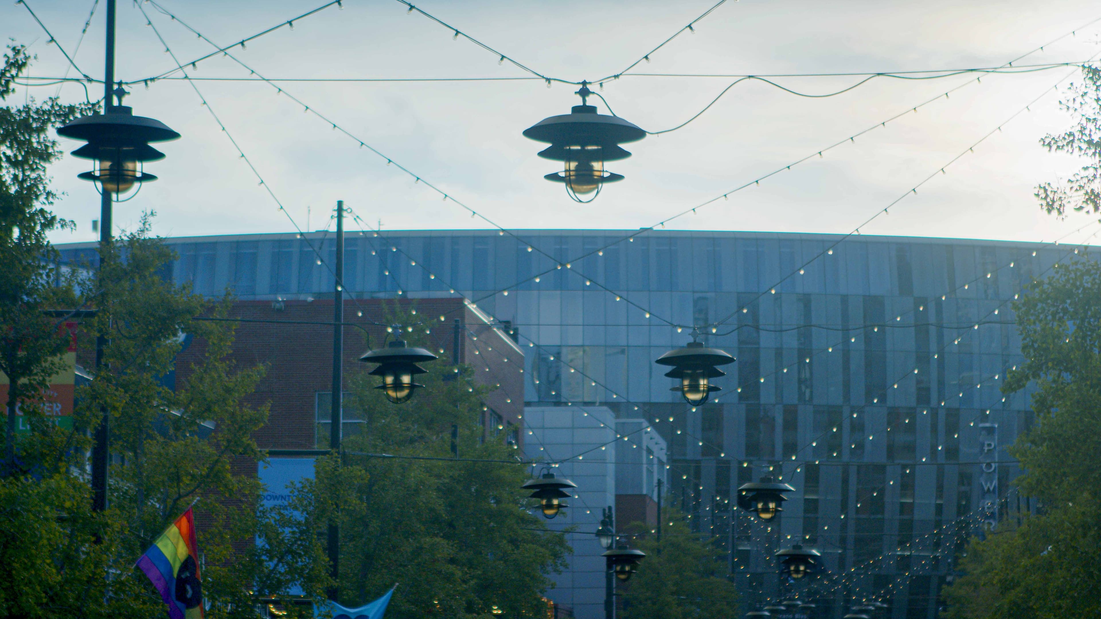 String lights hang above a street lined with trees, with a modern building in the background.
