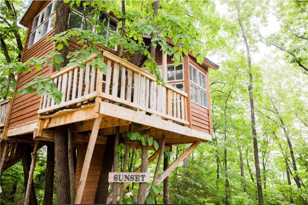A wooden treehouse perched among lush green trees with a sign that reads "SUNSET."