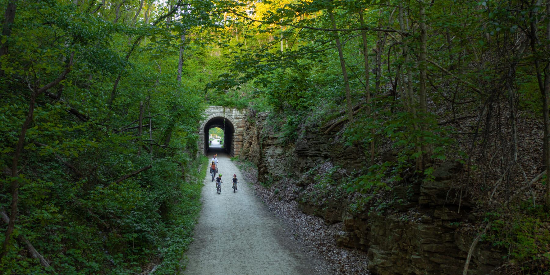 Cyclists ride along a forested trail leading to a stone tunnel entrance.