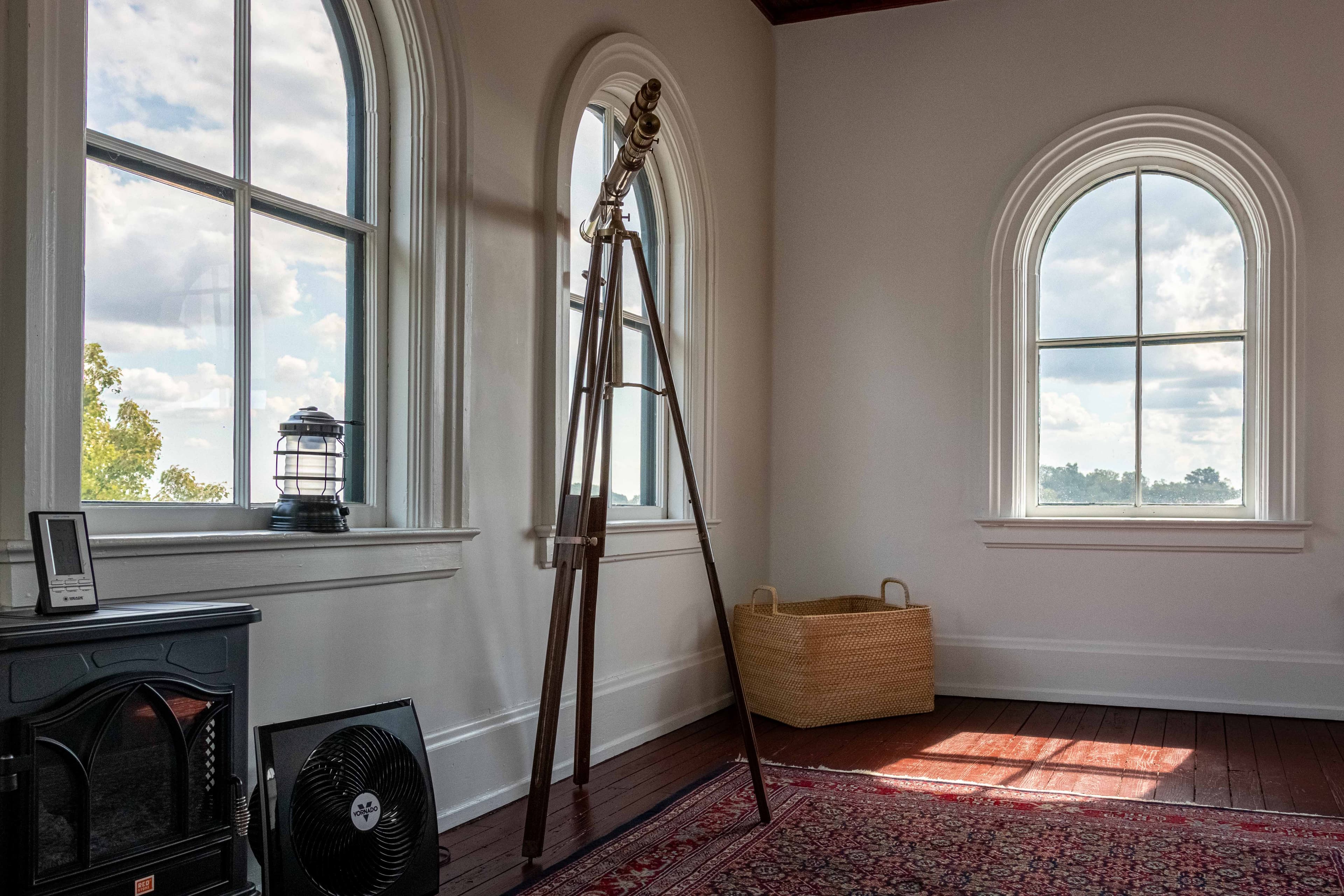 A cozy room featuring a telescope, a vintage lantern, a fan, and a wicker basket near large arched windows.