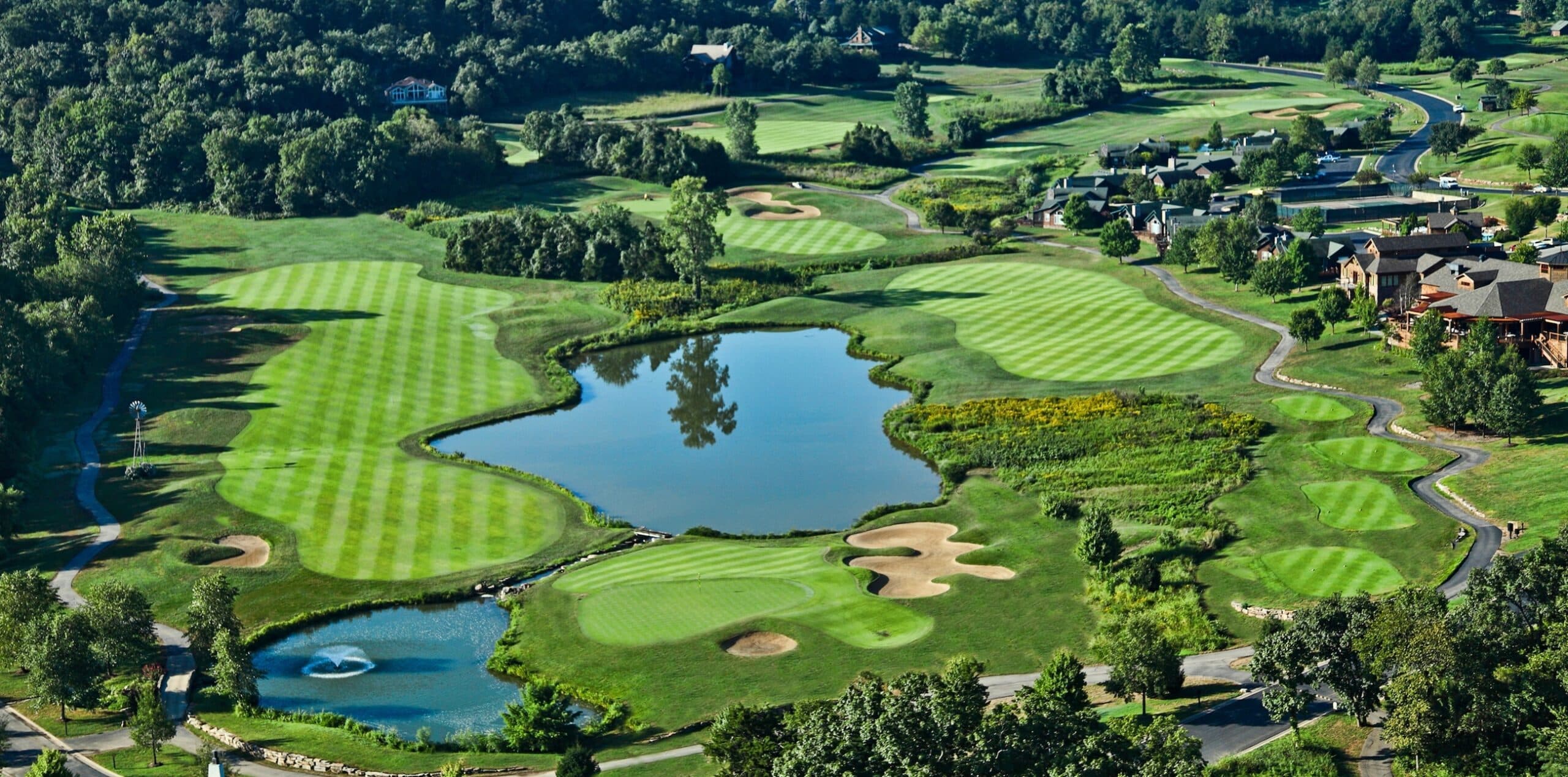 Aerial view of a golf course featuring lush green fairways, bunkers, and a pond surrounded by trees and homes.