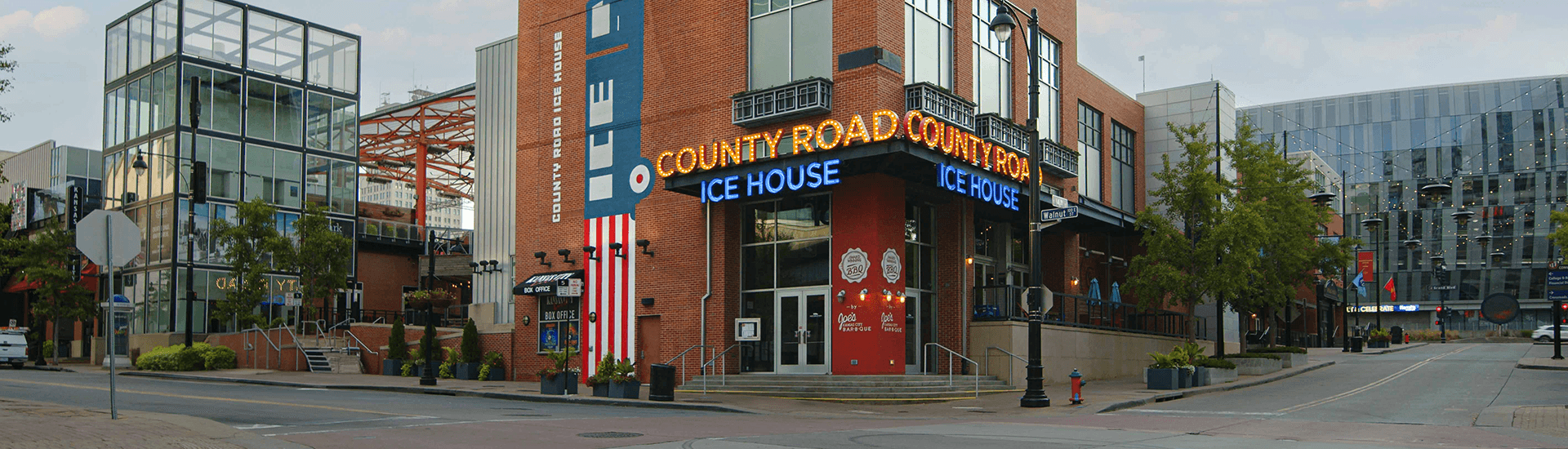 A modern brick building with a large sign reading "County Road Ice House" is seen on a city street lined with trees and other shops.