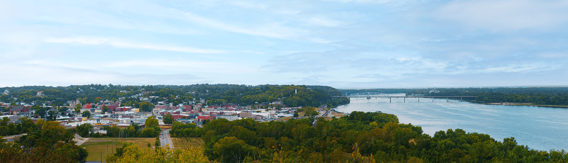 Panoramic view of a river town surrounded by green hills under a cloudy sky.